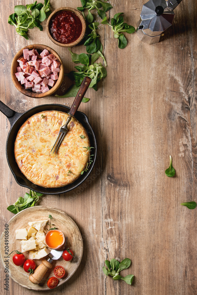 Spanish potato omelette tortilla with bacon served in cast-iron pan with sauce and ingredients above over wooden background. Top view, space.