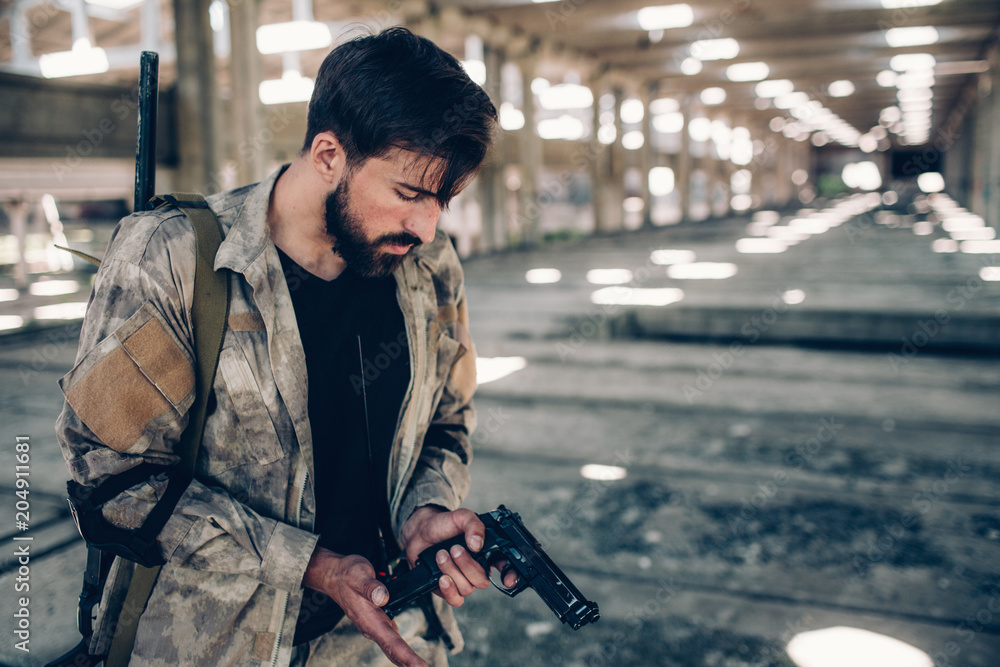 Serious professional is recharging his gun. He is holding his right hand at the low edge of pistol. Guy is looking at it. Young man is in big hangar. Serious professional is recharging his gun. He is holding his right hand at the low edge of pistol. Guy is looking at it. Young man is in big hangar.