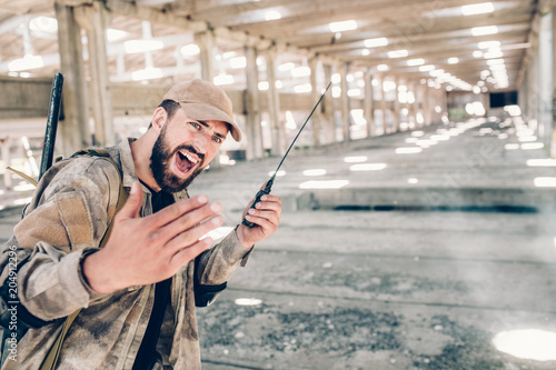 A picture of lonely bearded man in hangar. He is holding portable radio in left hand and using it. Guy is screaming. Also young man is keeping his right hand fingers together. photo