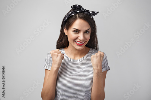 Picture of happy excited young female smiling broadly, making winning gesture with clenched fists after she won car in lottery. People, positive emotions, luck, triumph, success and achievement