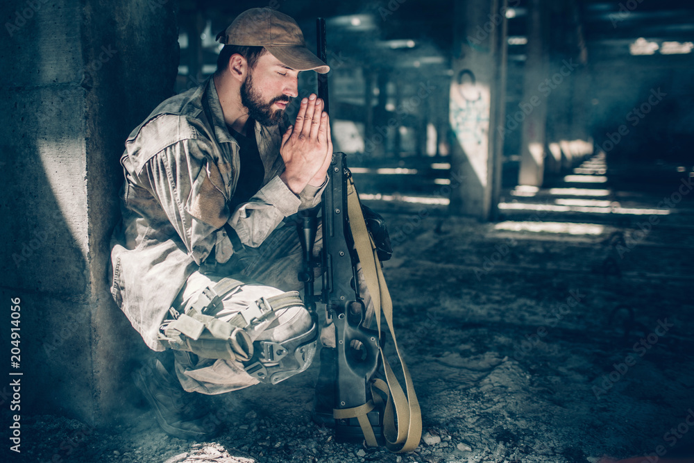 Spiritual man is sitting in squat position and praying. He is keeping his eyes closed and holding hands together close to face. Also there is a rifle near his knees.