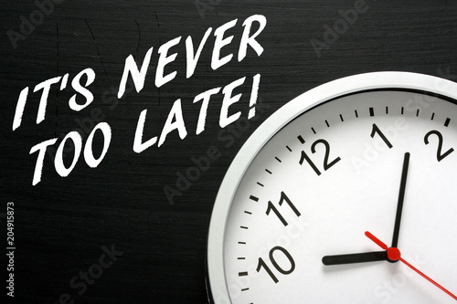 The phrase Its Never Too Late in white text on a blackboard next to a modern wall clock as a reminder to take action and avoid procrastination