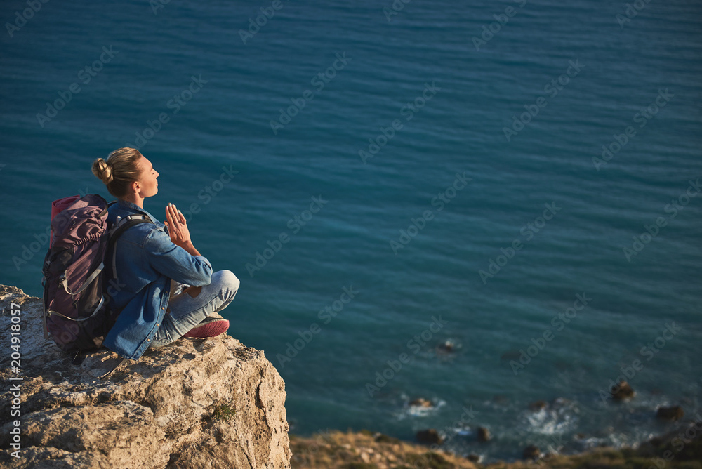 Peaceful girl with rucksack meditating on nature. She is holding palms together in front of chest. Copy space in right side