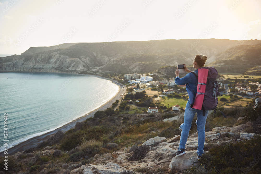 Back of girl in denim clothing taking picture of coastline from high. She is carrying backpack behind. Copy space in left side
