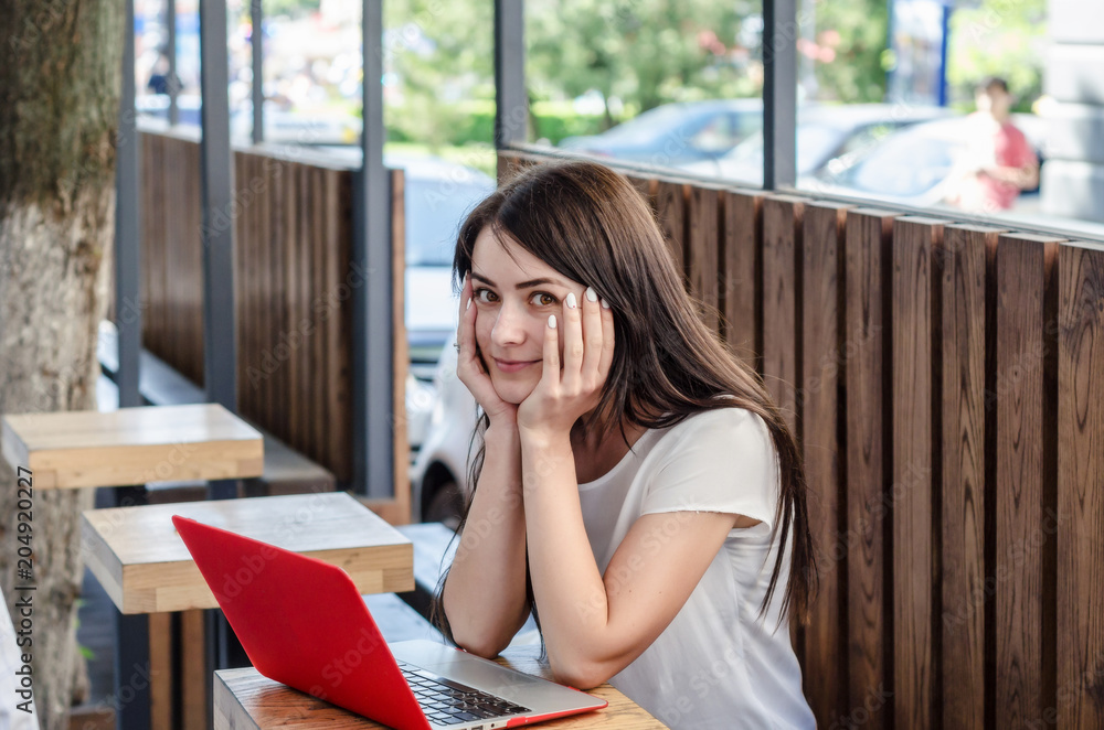 Beautiful girl sitting at a table in a street cafe with a red laptop