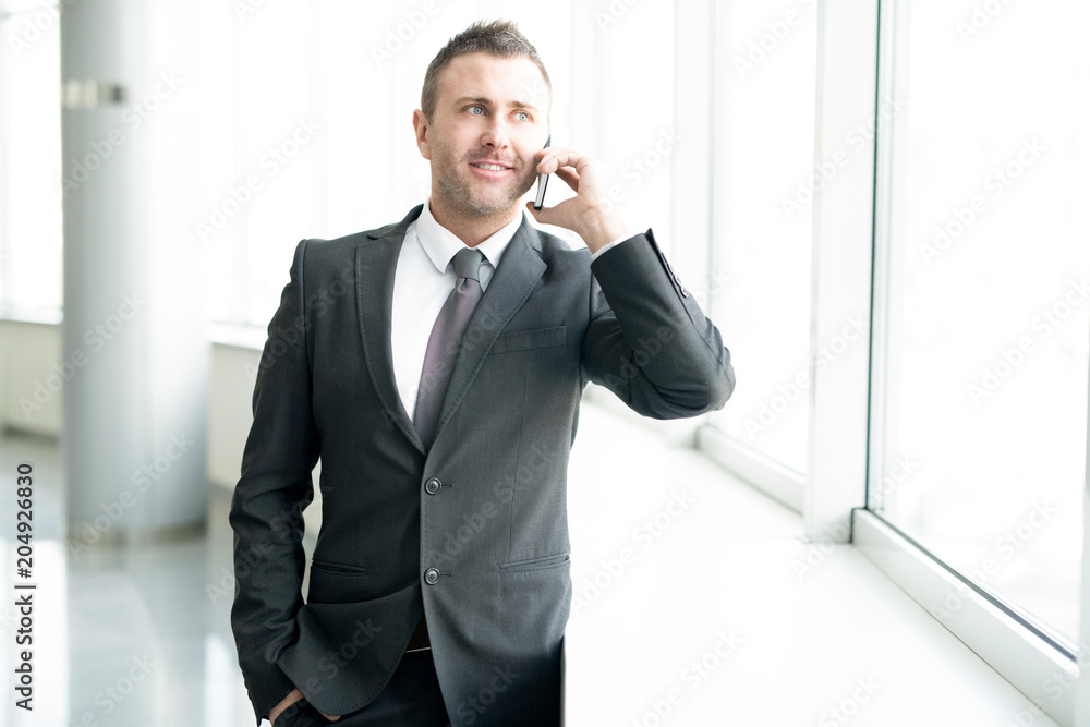 Waist up portrait of successful businessman standing by window in office and speaking by phone smiling cheerfully, copy space Waist up portrait of successful businessman standing by window in office and speaking by phone smiling cheerfully, copy space