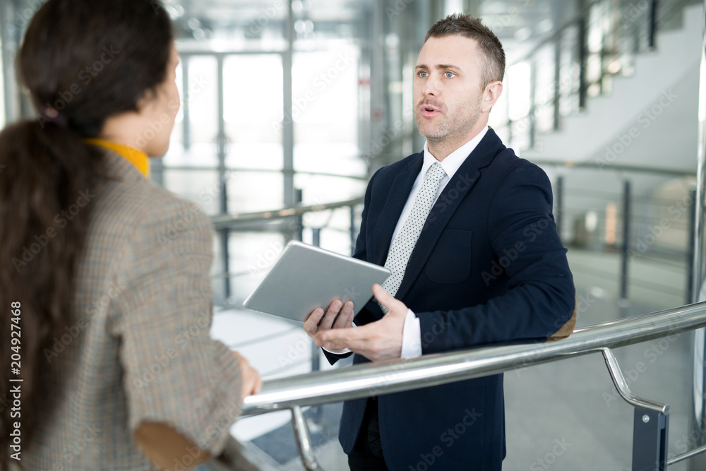 Waist up portrait of modern businessman holding laptop talking to female colleague standing by railing in office building, copy space Waist up portrait of modern businessman holding laptop talking to female colleague standing by railing in office building, copy space