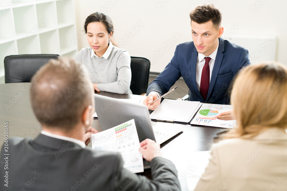 High angle view at business meeting with handsome young man explaining partnership terms to colleagues sitting at table in conference room High angle view at business meeting with handsome young man explaining partnership terms to colleagues sitting at table in conference room