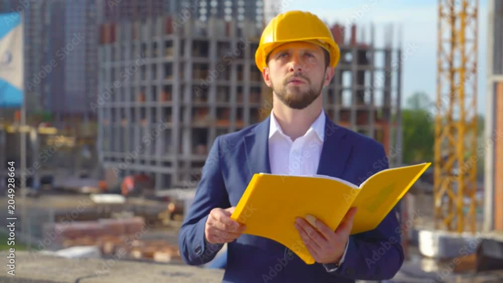 young attractive man in a yellow hard hat looks into the camera. Portrait of a handsome business man. the builder makes notes in a yellow folder. high-rise buildings under construction in the young attractive man in a yellow hard hat looks into the camera. Portrait of a handsome business man. the builder makes notes in a yellow folder. high-rise buildings under construction in the