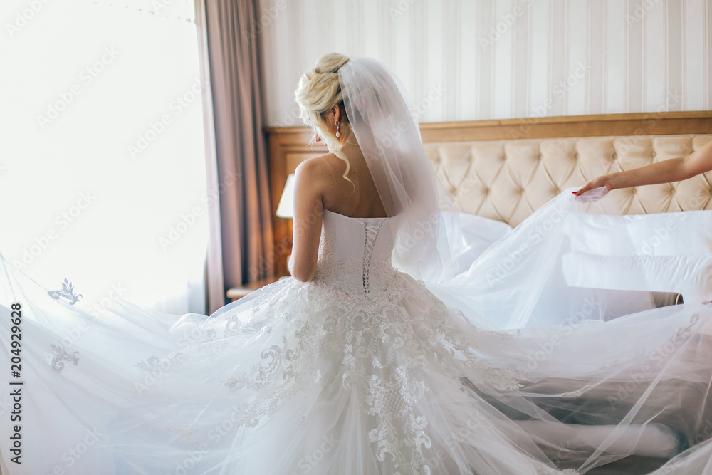 Bride with bridesmaids posing in hotel or fitting room at wedding day. Bridesmaids help bride to wear wedding dress. long plume in the dress
