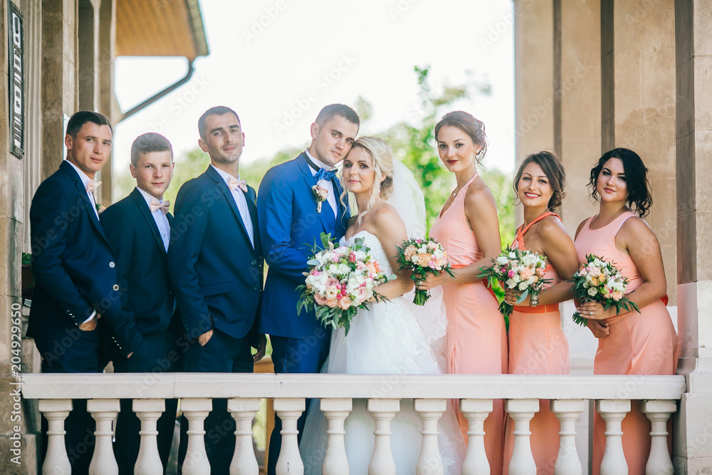 Cheerful & fun groom with bride, bridesmaids & groomsmen posing outdoors. Newlywed couple with best friends make funny photo near old castle.