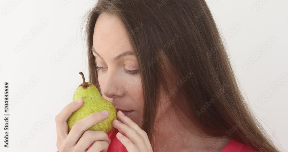 Young woman is eating a big juicy pear at white background. Girl takes first bite and then offer bite to viewer and saying Wanna bite