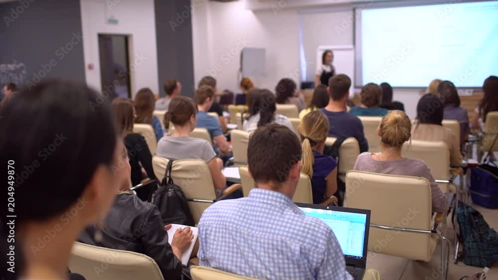 Education classroom blur background of university students sitting in a lecture hall or seminar room with teacher. View from behind audience at business conference looking towards female speaker Education classroom blur background of university students sitting in a lecture hall or seminar room with teacher. View from behind audience at business conference looking towards female speaker