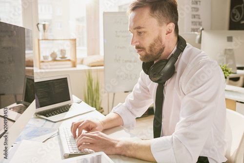 Side view portrait of handsome bearded businessman wearing shirt and tie sitting at desk in office and using modern computer, typing, with headphones on his neck