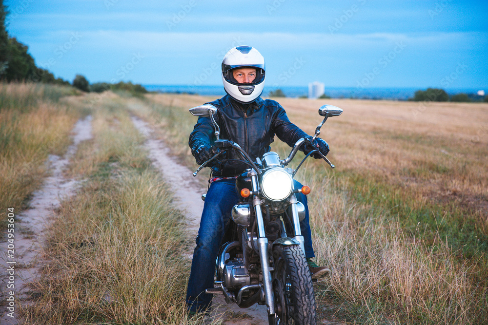 © lkoimages - Young biker wears black Leather Jacket and jeans, sits on his black old motorbike, holds white helmet and looks on beautiful soft sunset