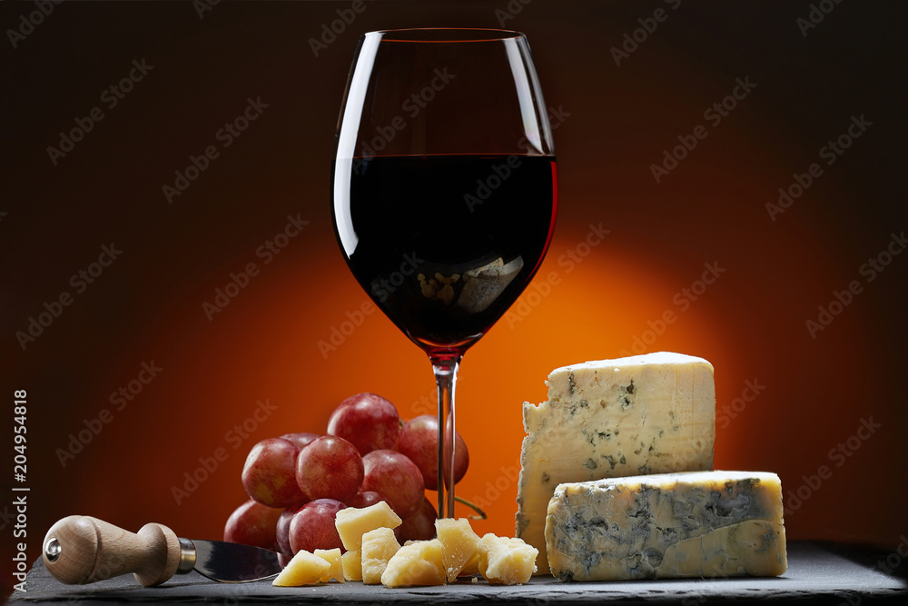 Glass of wine with grapes and a piece of cheese with mold, Parmesan cheese and cheese knife.  Orange background.