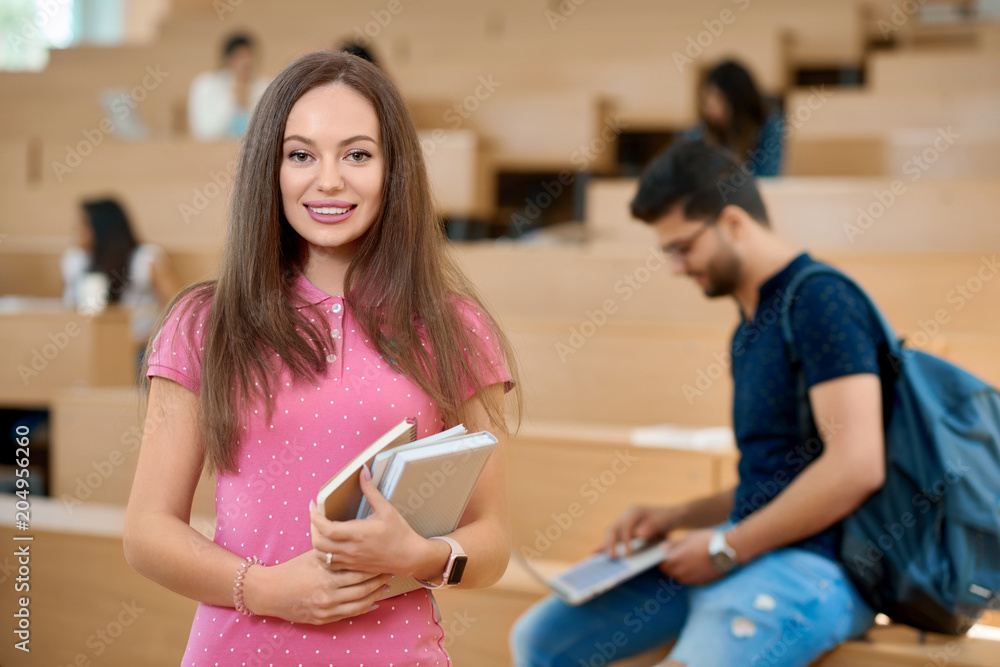 Pretty student keeping books in the modern spacy university classroom with wooden cascade desks. Standing on her groupmates background. Prepearing for exams. Smiling, feeling satisfied. Pretty student keeping books in the modern spacy university classroom with wooden cascade desks. Standing on her groupmates background. Prepearing for exams. Smiling, feeling satisfied.