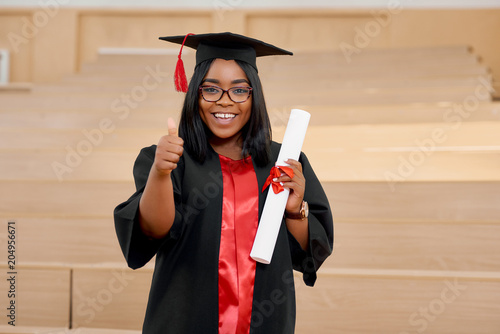 Positive girl graduating from university. Student wearing black and red education gown and keeping diploma.Standing in classroom with wooden cascade desks. Smiling, feeling happy.