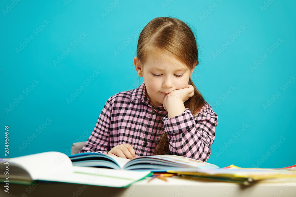Disciplined girl attentively reads book sitting at the table, expresses her passion, on isolated blue background. Child performs home task. Disciplined girl attentively reads book sitting at the table, expresses her passion, on isolated blue background. Child performs home task.