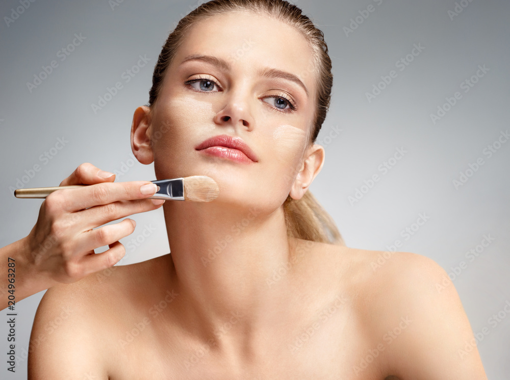 Beautiful woman applying liquid foundation on her face on grey background Beautiful woman applying liquid foundation on her face on grey background