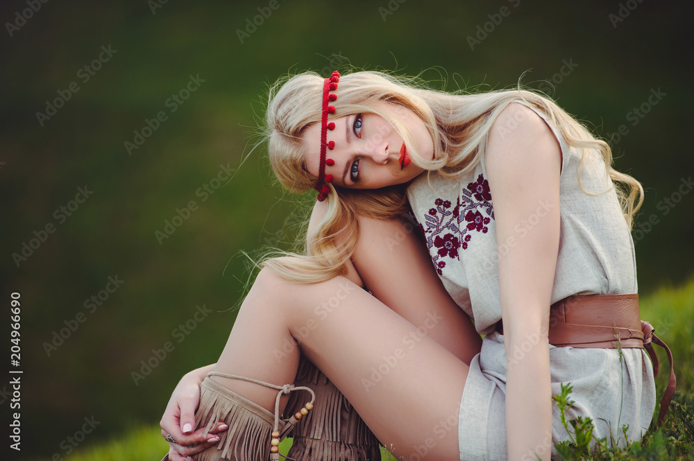 Naklejka premium tender blonde woman sits on the grass dressed in a natural embroidered dress bandage on her head, on a green background stylish and cozy. The concept of advertising the love of nature. place for text