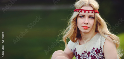 tender blonde woman sits on the grass dressed in a natural embroidered dress bandage on her head, on a green background stylish and cozy. The concept of love advertising is panoramic. place for text