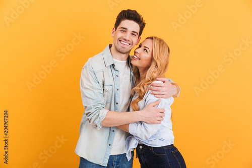 Portrait of young lovely people man and woman in basic clothing hugging together with happy smile, isolated over yellow background