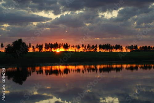 Beautiful orange sunset shines through the trees on the river in the summer against the blue sky with evening clouds reflected in the smooth water