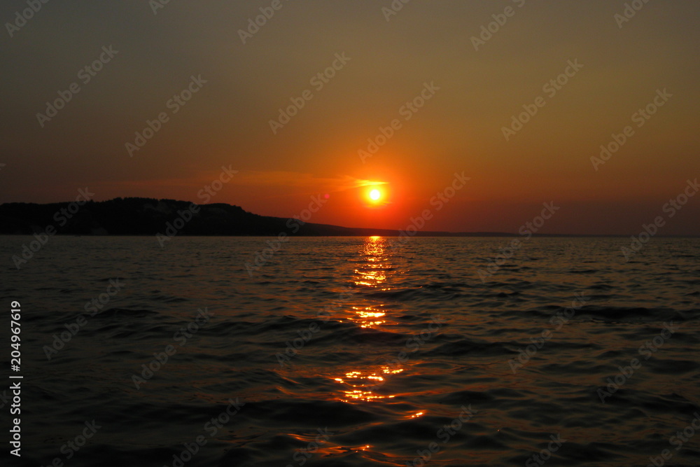 Beautiful orange sunset on the lake in summer against the setting sun on the horizon and the evening cloudless sky above the water