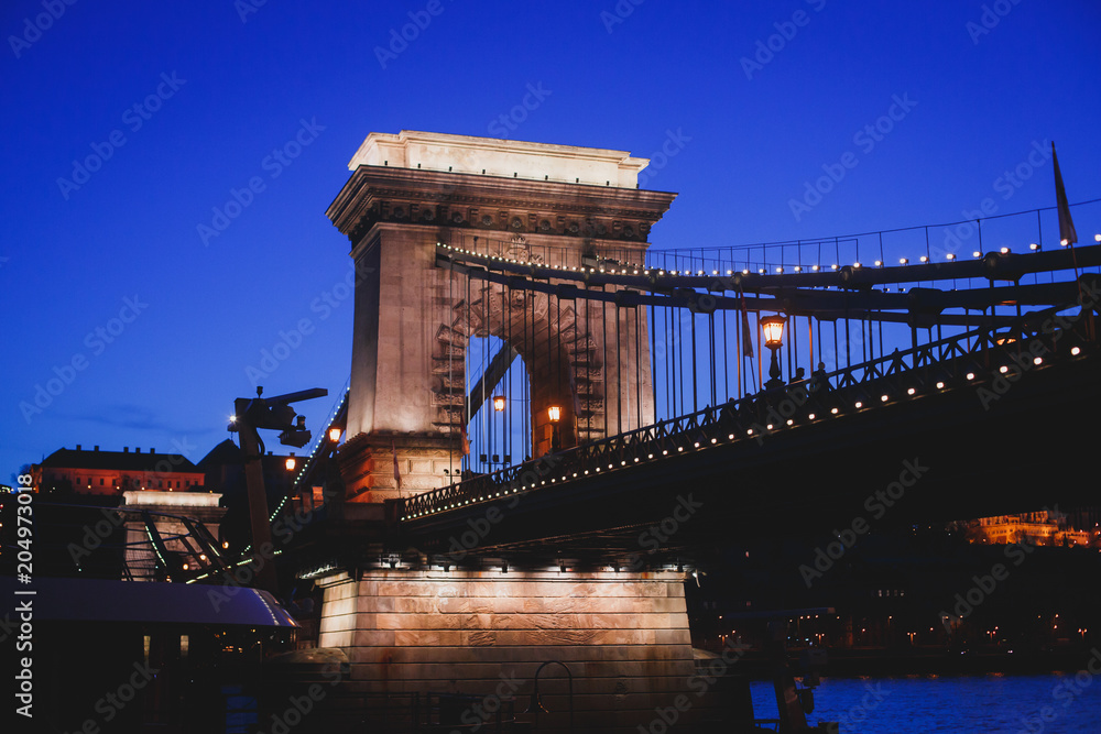 Obraz premium Night view of a famous Budapest Szechenyi Chain Bridge, a suspension bridge that spans the River Danube between Buda and Pest, the western and eastern sides of Budapest