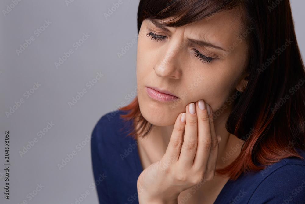 Closeup portrait of young woman on grey background suffering from toothache, holding fingers to jaw and closing eyes from pain. Closeup portrait of young woman on grey background suffering from toothache, holding fingers to jaw and closing eyes from pain.