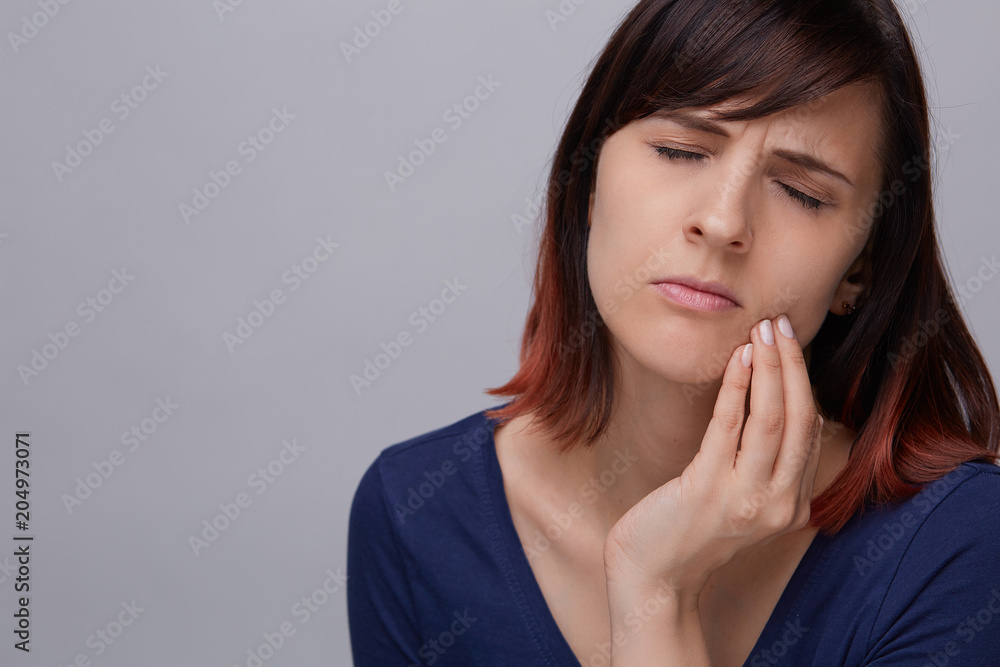 Closeup portrait of young woman on grey background suffering from toothache, holding fingers to jaw and closing eyes from pain. Closeup portrait of young woman on grey background suffering from toothache, holding fingers to jaw and closing eyes from pain.