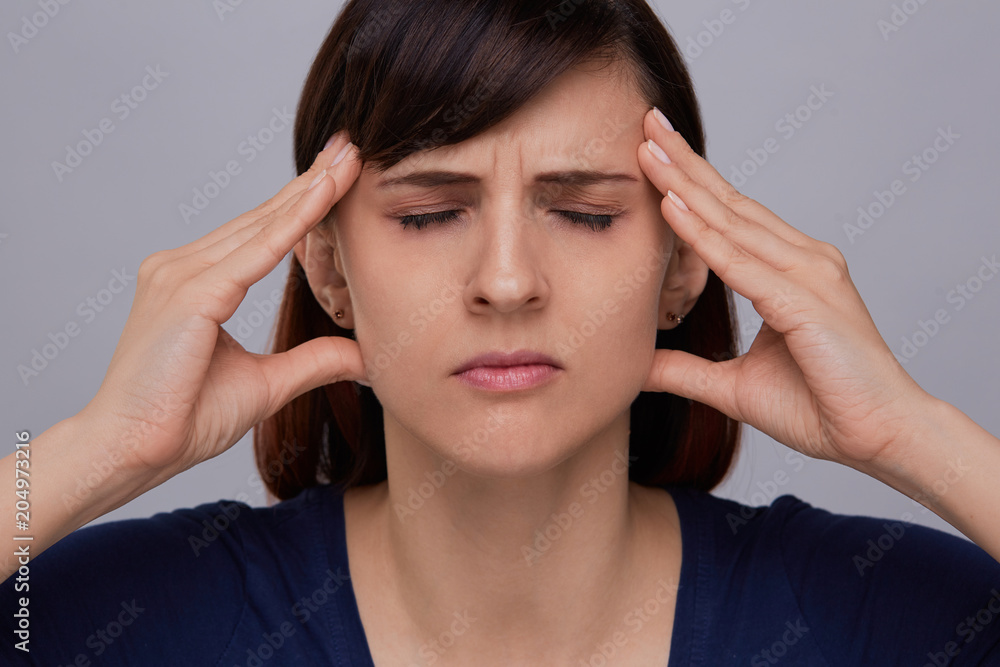 Closeup portrait of young woman on grey background suffering from strong headache, holding fingers to temples and closing eyes from pain.Severe migraine. Closeup portrait of young woman on grey background suffering from strong headache, holding fingers to temples and closing eyes from pain.Severe migraine.