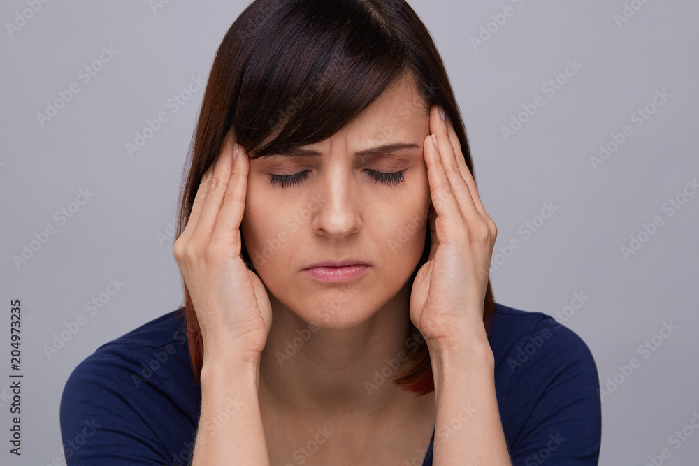 Closeup portrait of young woman on grey background suffering from strong headache, holding fingers to temples and closing eyes from pain.Severe migraine. Closeup portrait of young woman on grey background suffering from strong headache, holding fingers to temples and closing eyes from pain.Severe migraine.