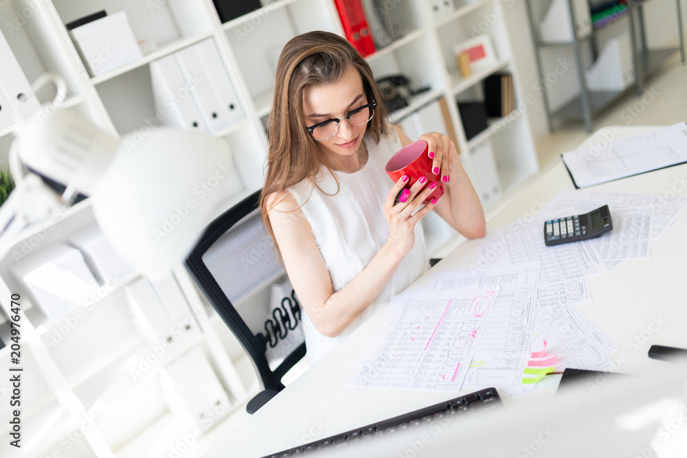 A young girl in the office holds a pink marker, a red mug and works with documents. A young girl in the office holds a pink marker, a red mug and works with documents.