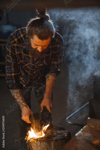 Blacksmith works as a hand hammer with molten metal on the anvil in the smithy with a spark firework.
