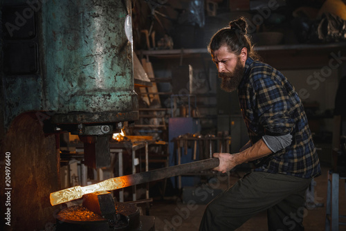 Brutal blacksmith works in his workshop on a mechanical hammer holding a metal part in his hands. Portrait of a profession.