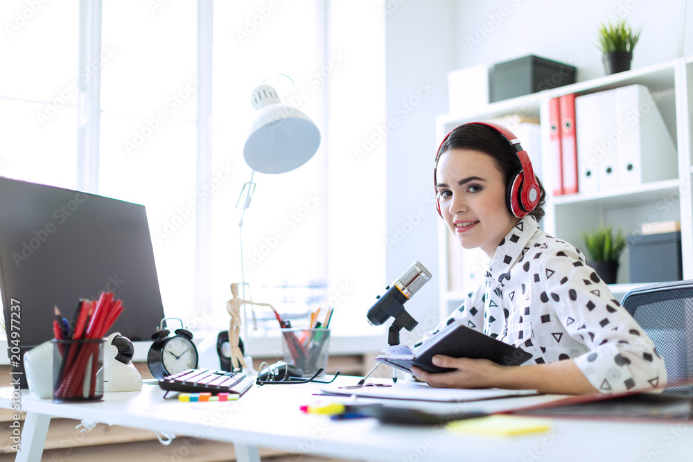 Beautiful young girl sits in headphones and with a notepad at the table in the office and speaks into the microphone. Beautiful young girl sits in headphones and with a notepad at the table in the office and speaks into the microphone.