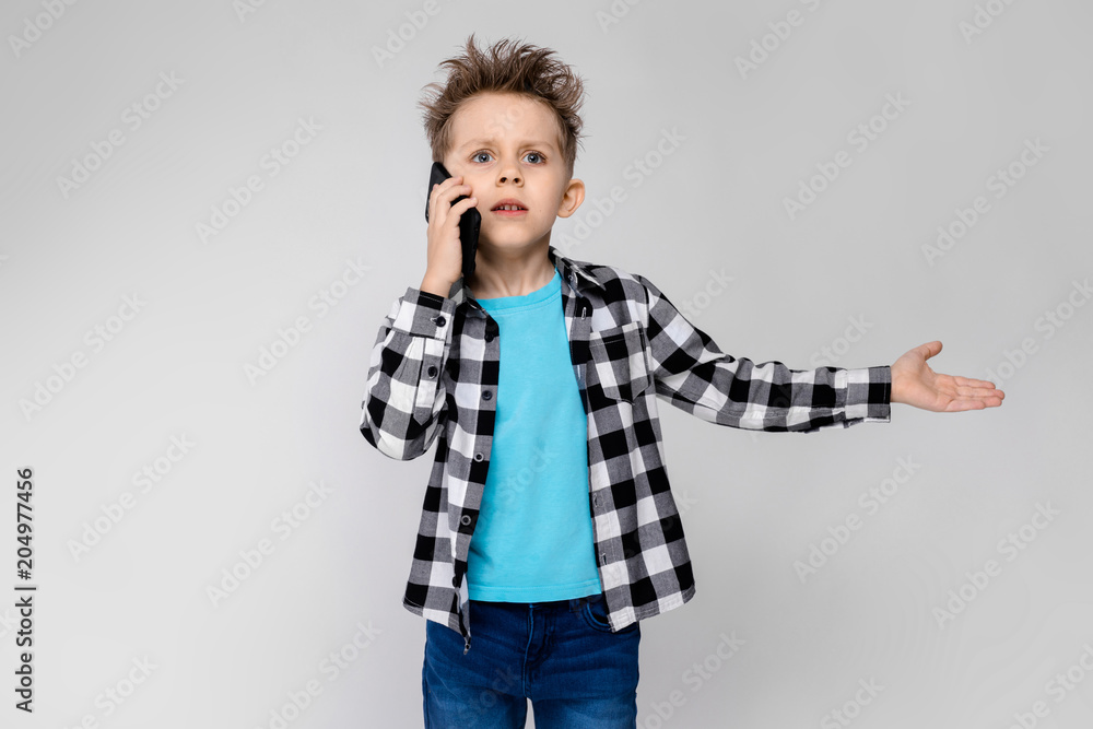 A handsome boy in a plaid shirt, blue shirt and jeans stands on a gray background. The boy is holding a phone A handsome boy in a plaid shirt, blue shirt and jeans stands on a gray background. The boy is holding a phone