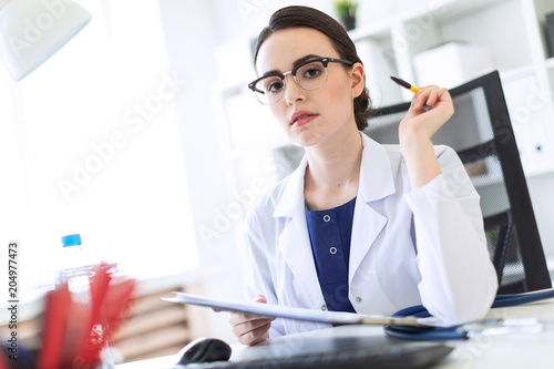 A beautiful young girl in a white robe is sitting at a computer desk with documents and a pen in her hands. Photo with depth of field, focus on girl. photo