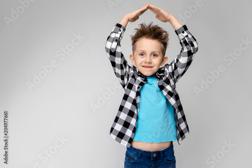 A handsome boy in a plaid shirt, blue shirt and jeans stands on a gray background. The boy folded his arms over his head in the form of a house