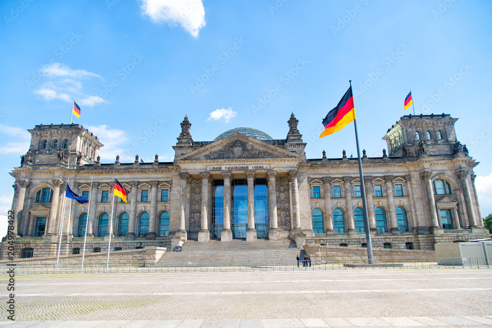 Naklejka premium German flags waving in the wind at famous Reichstag building, seat of the German Parliament (Deutscher Bundestag), on a sunny day with blue sky and clouds, central Berlin Mitte district, Germany