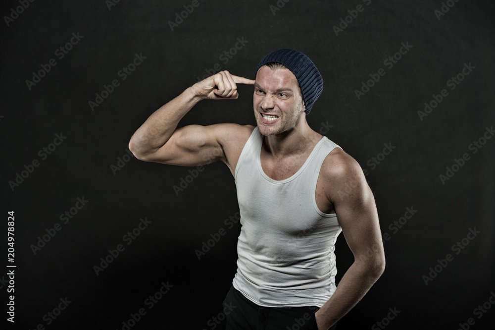 Foto de Man with arms pointing at head with index finger. Masculinity concept. Macho on aggressive face with strong muscles look brutal, black background. Man in hat and sleeveless undershirt