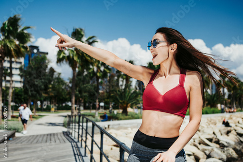 Look there. Joyful young woman is pointing finger up with excitement. She is relaxing on the seaside bridge in tropical place