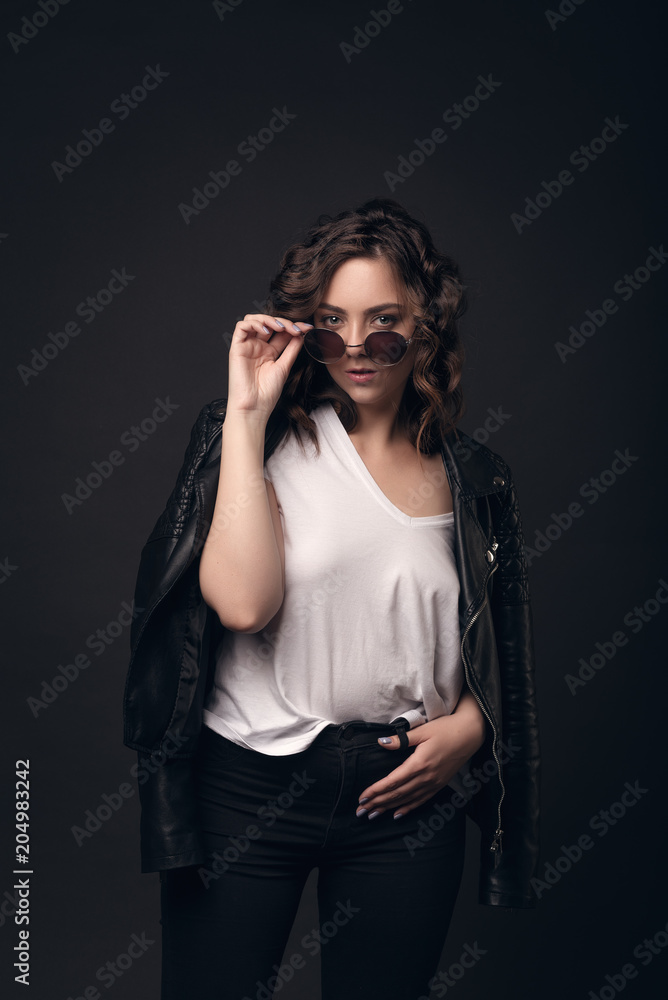 A young brunette girl with a stylish hairstyle on a dark background in a leather jacket, white T-shirt and sunglasses. Professional model posing on an isolated dark background with space for text A young brunette girl with a stylish hairstyle on a dark background in a leather jacket, white T-shirt and sunglasses. Professional model posing on an isolated dark background with space for text