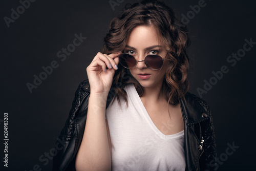 A young brunette girl with a stylish hairstyle on a dark background in a leather jacket, white T-shirt and sunglasses. Professional model posing on an isolated dark background with space for text