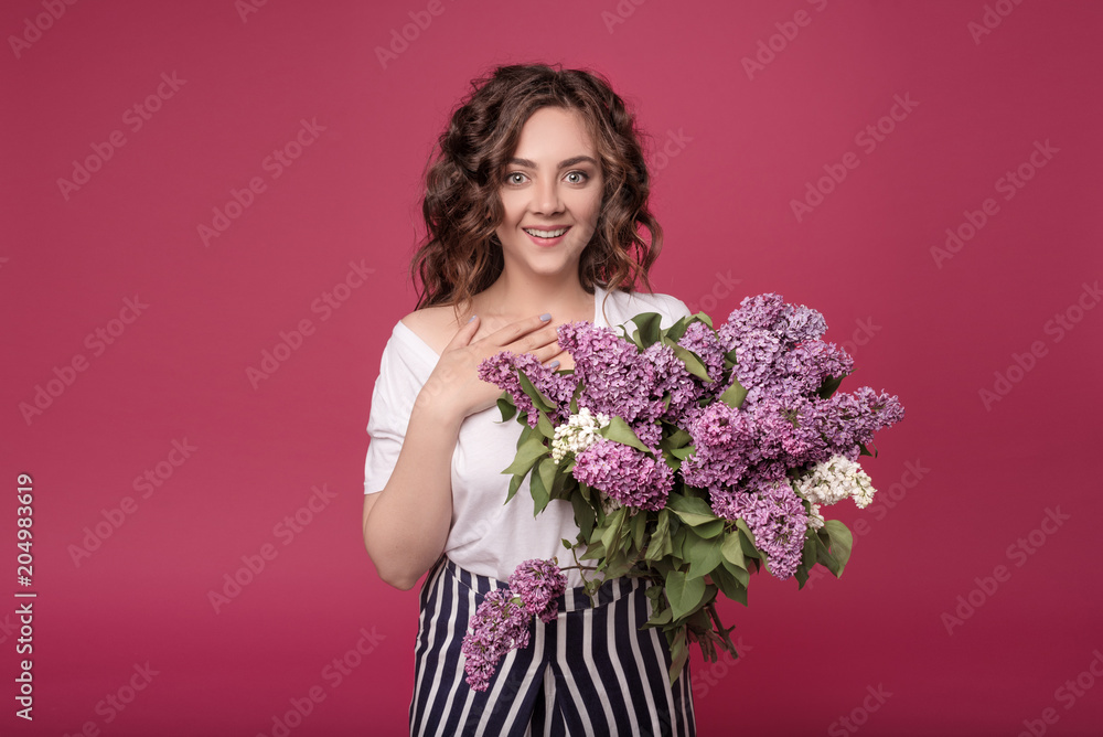 A young slender girl in stylish clothes is holding a bouquet of purple flowers. She is surprised at the gift she was given on a date. On a pink background with space for text