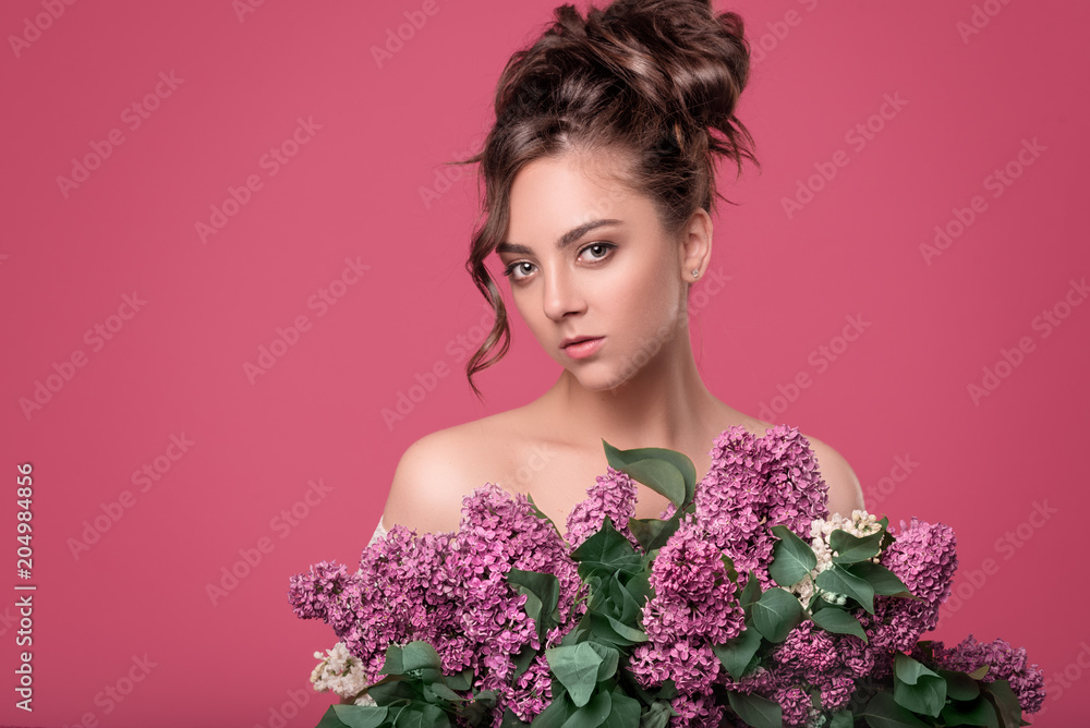 Young cute tender girl model with curly hair and professional make-up holds in hands a beautiful bouquet of purple flowers on a pink isolated background with space for text Young cute tender girl model with curly hair and professional make-up holds in hands a beautiful bouquet of purple flowers on a pink isolated background with space for text