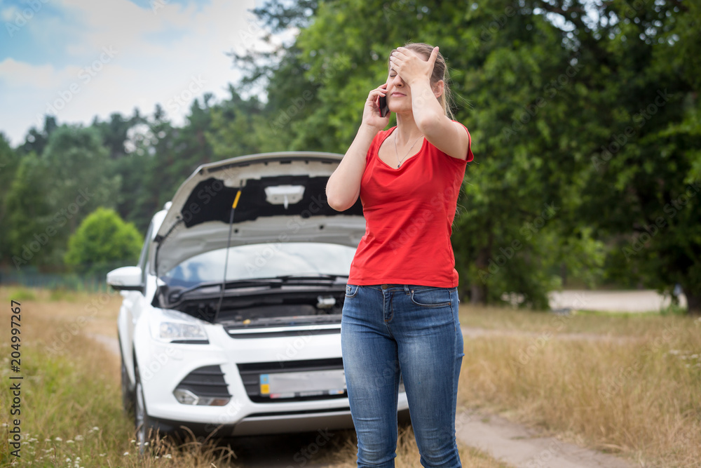 Upset young woman standing at her broken car and calling car assitance