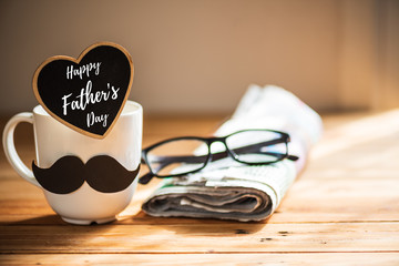 Happy fathers day concept. coffee cup with black paper mustache, heart tag with  Happy father's day text and newspaper, glasses on wooden table background.